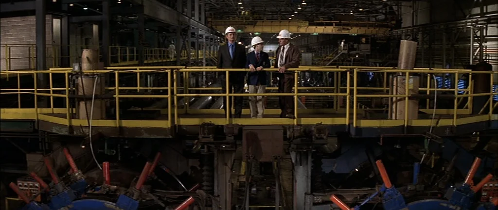 Rushmore (1998) movie still: wide — A vast industrial plant features a yellow-railed metal walkway suspended over heavy machinery. Three men wearing…