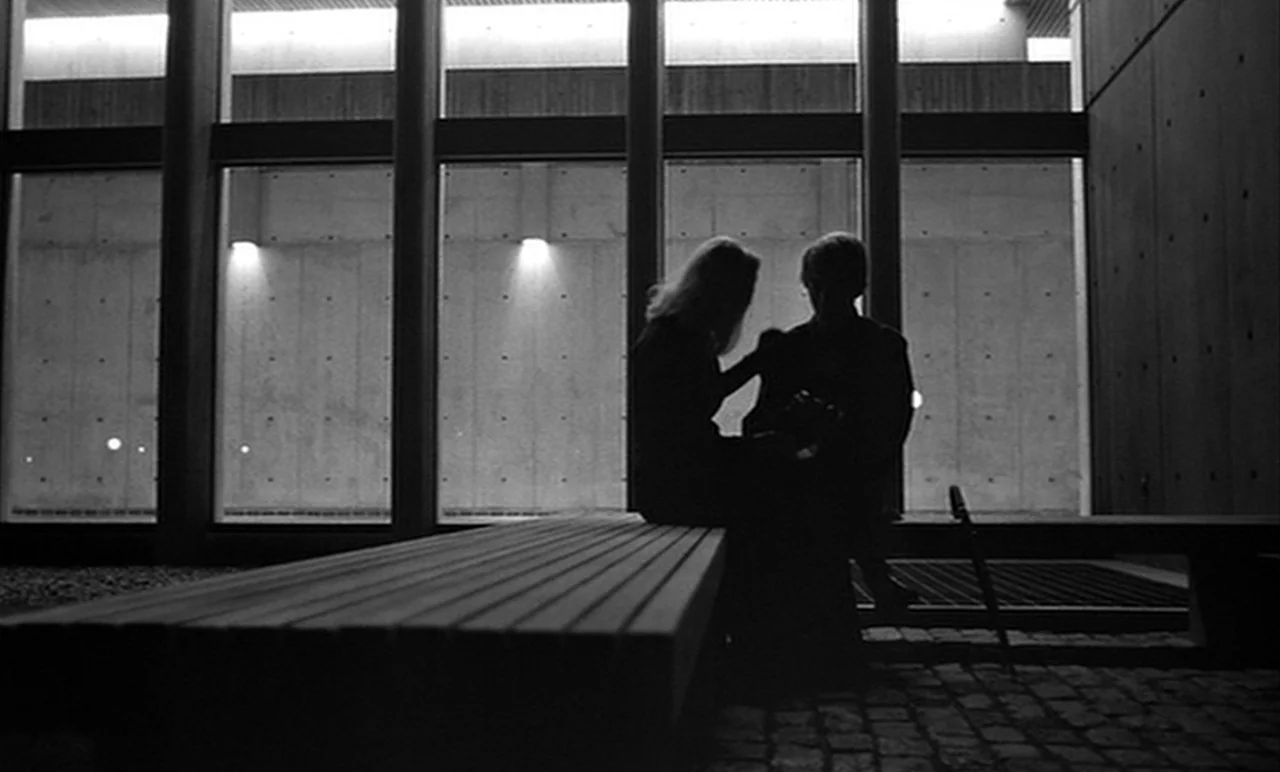 Stereo (1969) movie still: medium — A modern building lobby features floor-to-ceiling windows and concrete walls at night. Two people sit…