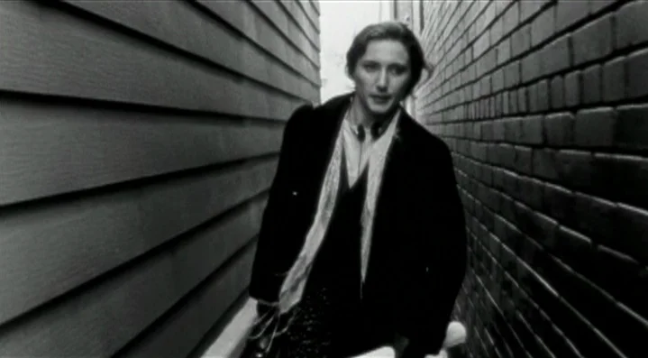 Roadkill (1989) movie still: medium — A person walks through a narrow exterior passage bordered by horizontal siding and brick. The subject moves…
