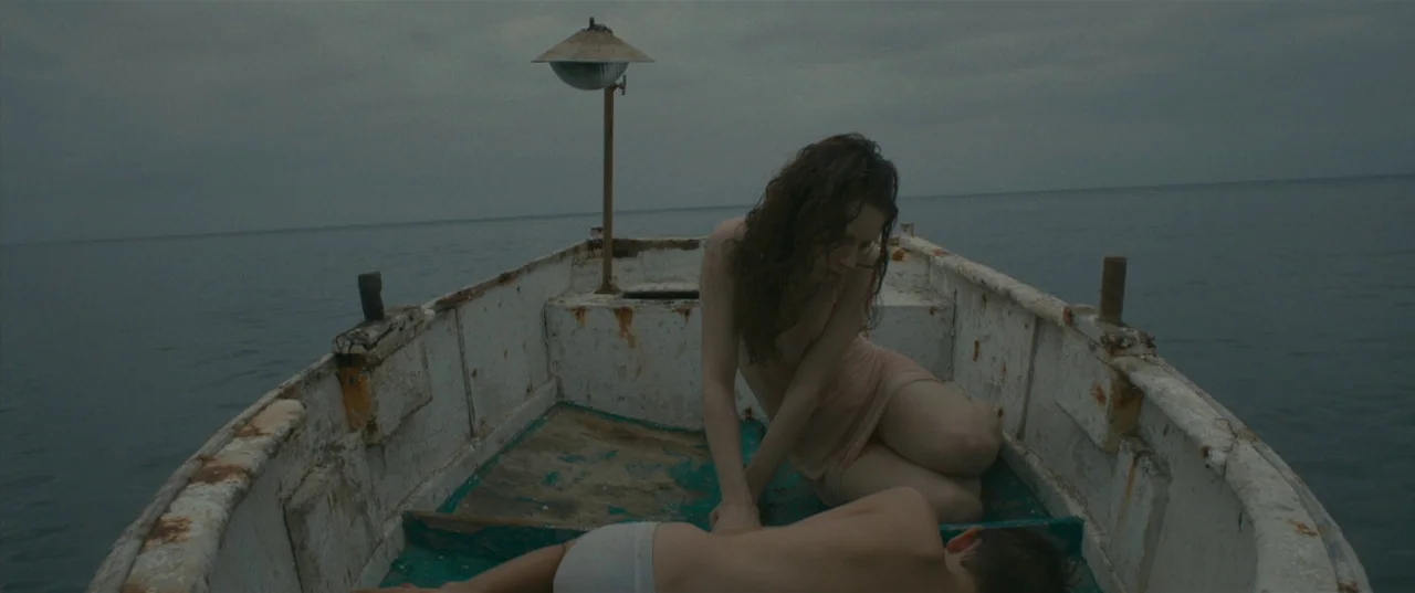 Evolution (2015) movie still: medium — A weathered wooden boat floats on a calm, overcast body of water. A woman leans over a child lying on the…