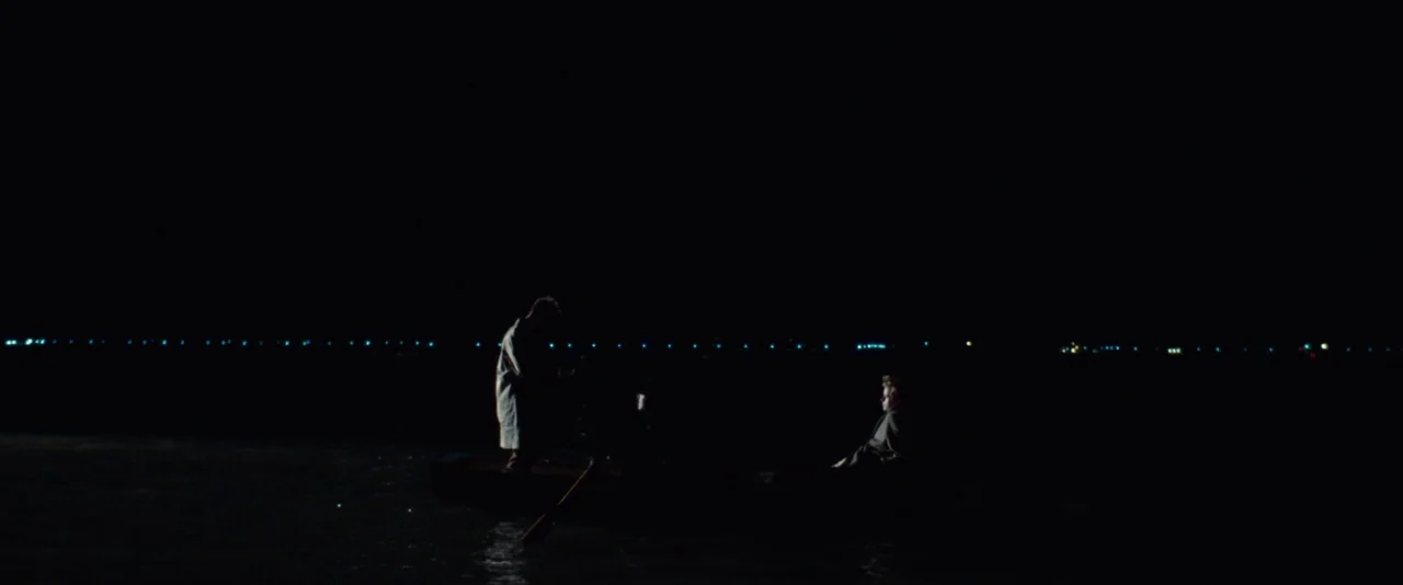 Kill Your Darlings (2013) movie still: wide — A small wooden rowboat sits on dark water under a vast, pitch-black night sky. Two figures in coats…
