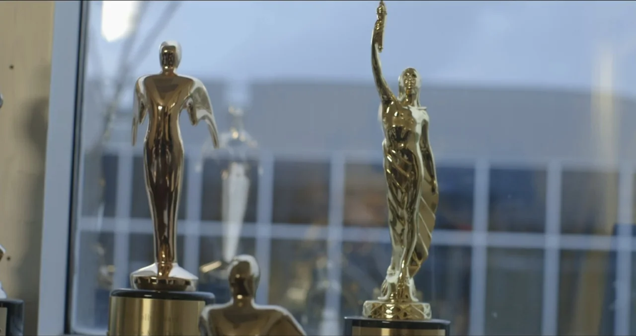 Faith, Love & Chocolate (2018) movie still: medium — Two metallic gold awards sit on a dark shelf before a large window. A view of a building…