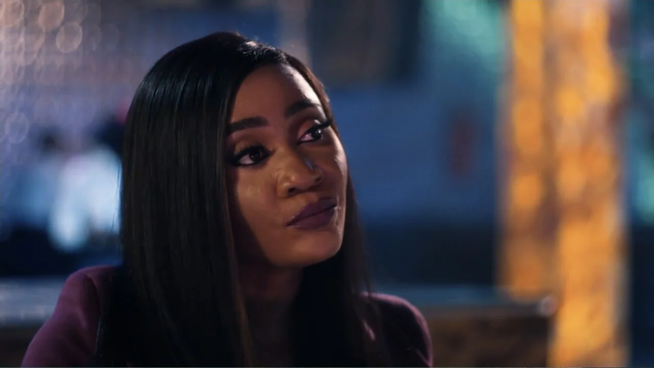 SIN: Blackout (2025) movie still: closeup — A dimly lit lounge setting with bokeh lights visible in the blurred background. A woman in a dark blazer…