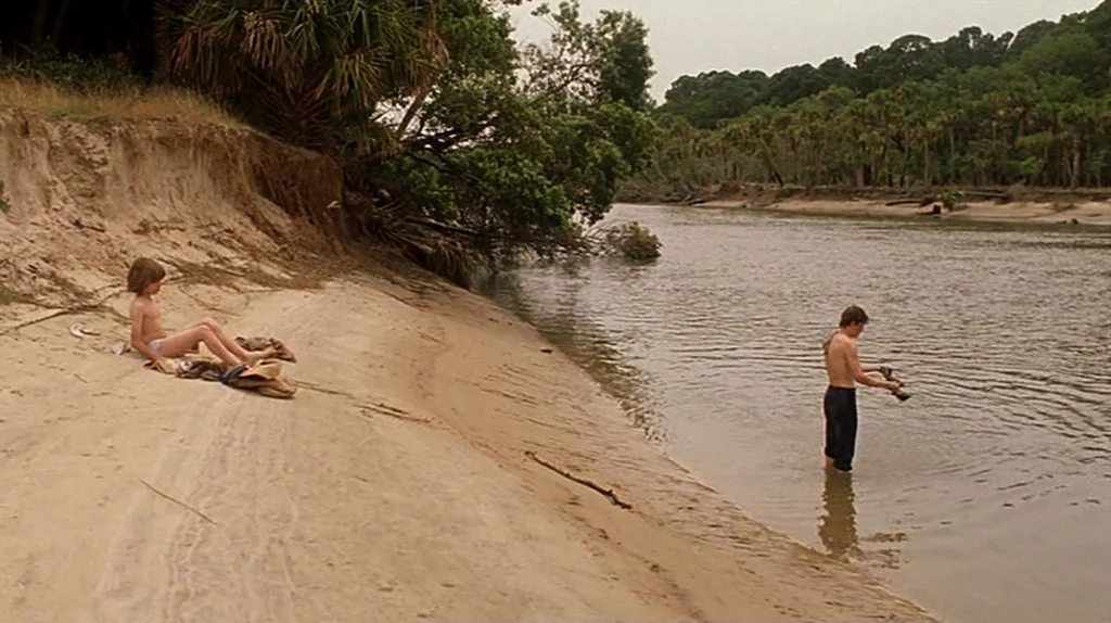 Undertow (2004) movie still: wide — A sandy riverbank is lined with dense forest and tropical foliage. Two young males are situated near the water,…