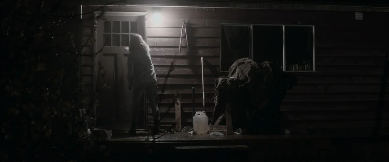 The Hollow Child (2018) movie still: fullbody — A dark wooden porch features a door and various tools leaning against the wall. A woman in a plaid…