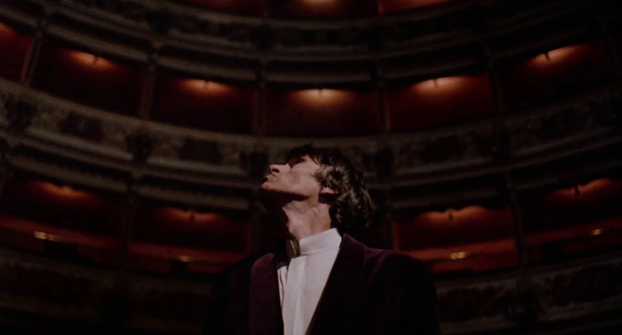 Etoile (1989) movie still: medium — The interior of a grand historic opera house with multiple tiers of red-upholstered balconies. A man in a dark…