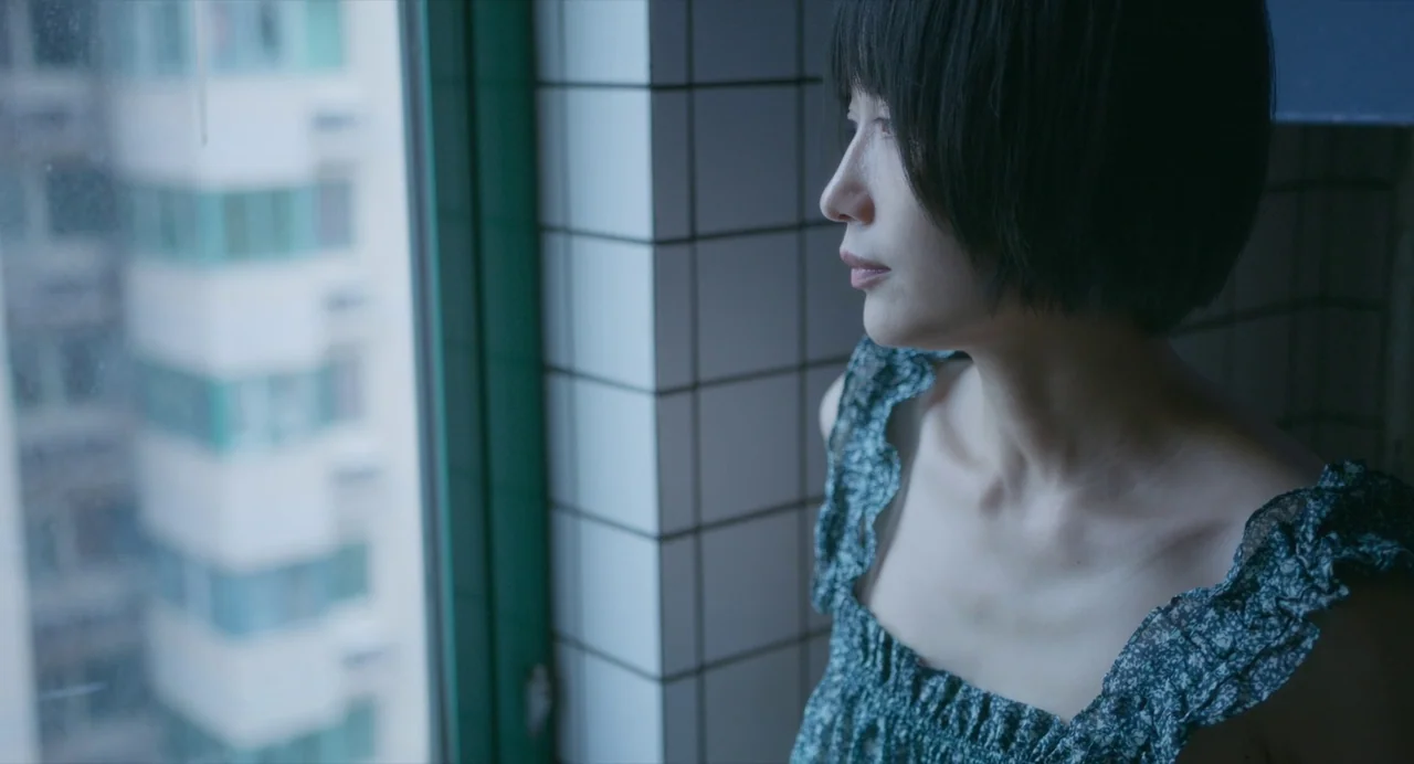 White River (2023) movie still: medium — A woman stands beside a window framed by white tiled walls in an interior space. She stares out of the…