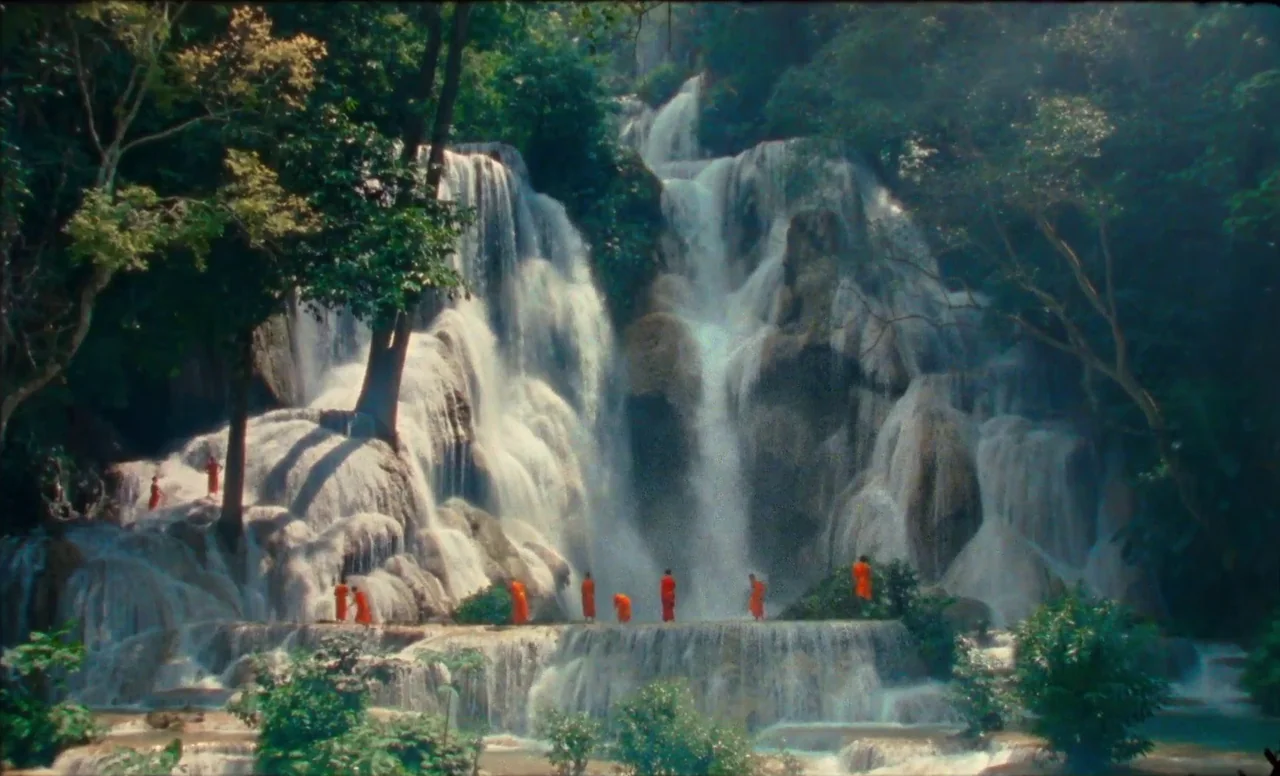 Samsara (2023) movie still: wide — A tiered limestone waterfall flows through a lush, green tropical forest environment. A group of monks in bright…
