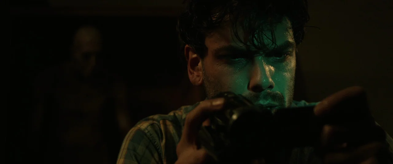 Terrified (2017) movie still: closeup — A man stands in a pitch-black interior space, his face illuminated by green light. He grips a small camera…