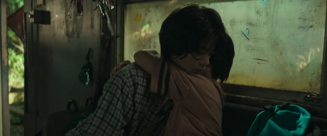 Monster (2023) movie still: medium — A dimly lit, weathered train interior features a window covered in grime and dust. Two teenagers sit side by…