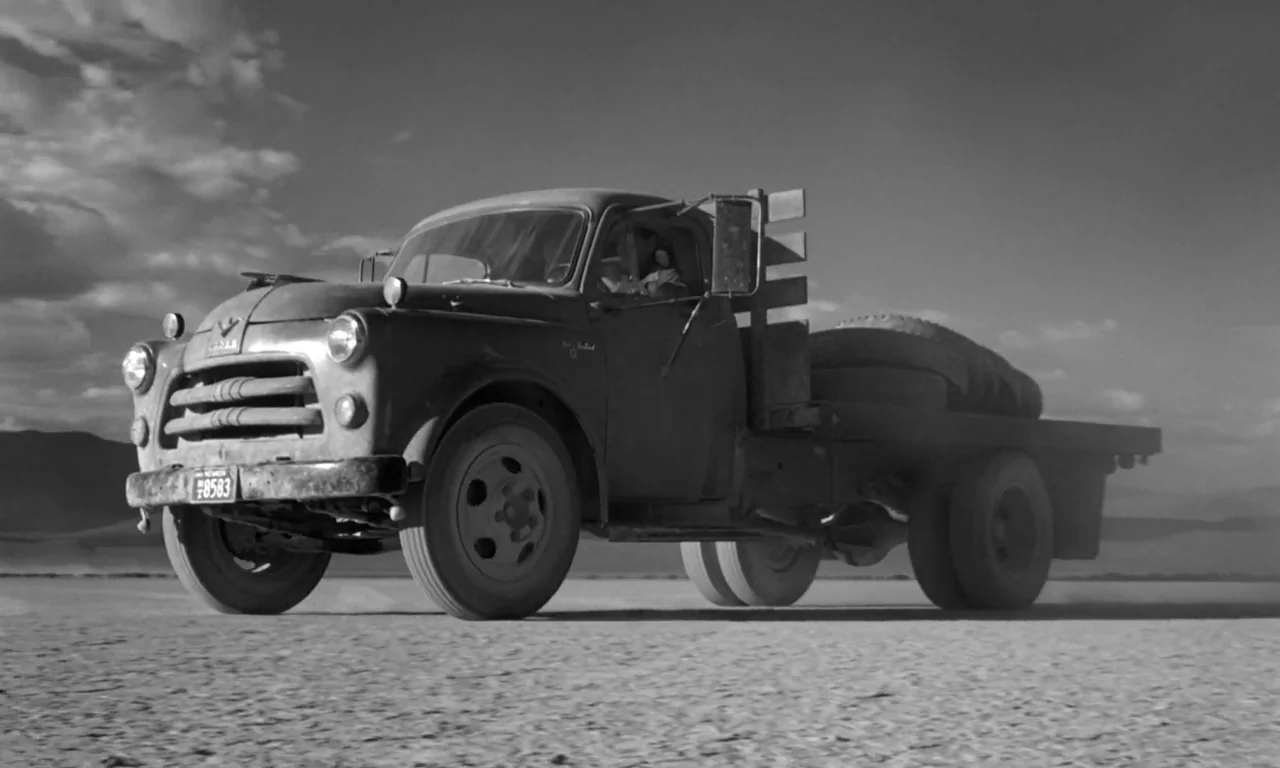 The Misfits (1961) movie still: fullbody — A vintage 1950s flatbed truck travels across a dusty, desolate dry lake bed. The driver operates the…