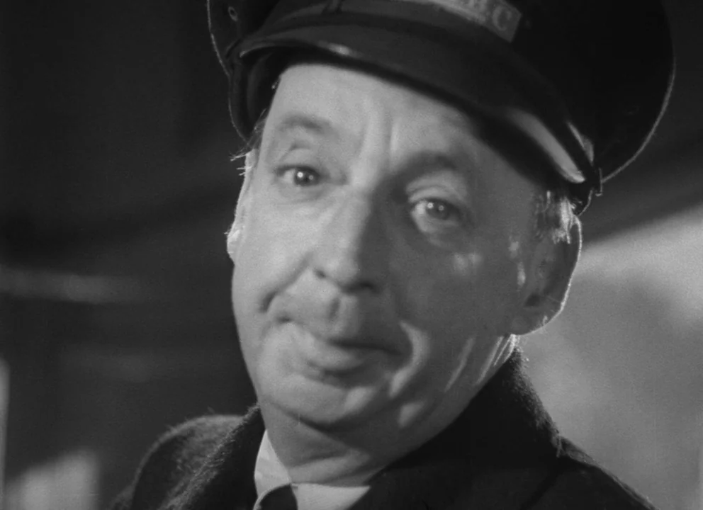 Dead of Night (1945) movie still: closeup — A dimly lit indoor space featuring a man wearing a dark service cap. The man gazes toward the lens with…