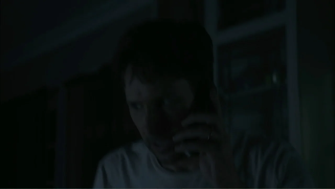 Dark Skies (2013) movie still: closeup — A dimly lit interior space with dark walls and minimal background detail. A man in a light t-shirt listens…