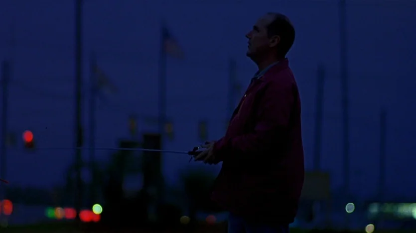 Love Liza (2002) movie still: medium — A man stands in a dark outdoor area at night with distant city lights. The man holds a remote control device…