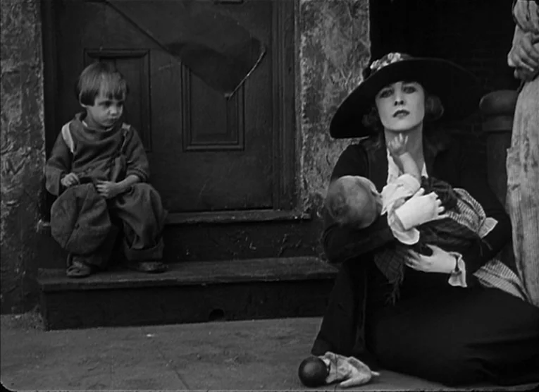 The Kid (1921) movie still: medium — A woman sits on the pavement before a dark wooden door with a small child. She cradles an infant in her arms…