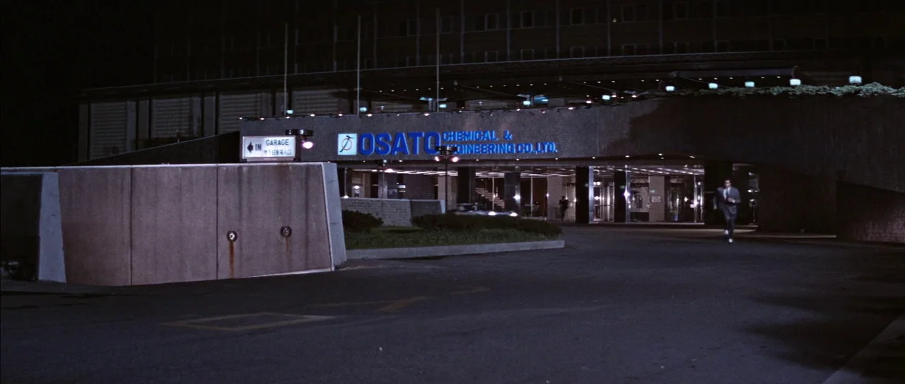 You Only Live Twice (1967) movie still: wide — A dark, paved entrance to a modern corporate building at night. A lone man in a suit runs across the…