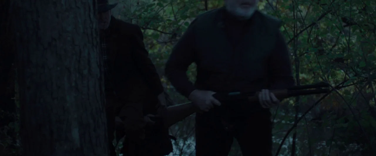 Wildling (2018) movie still: medium — Two men move through a dense, dark forest during the late hours of twilight. One man holds a rifle while the…