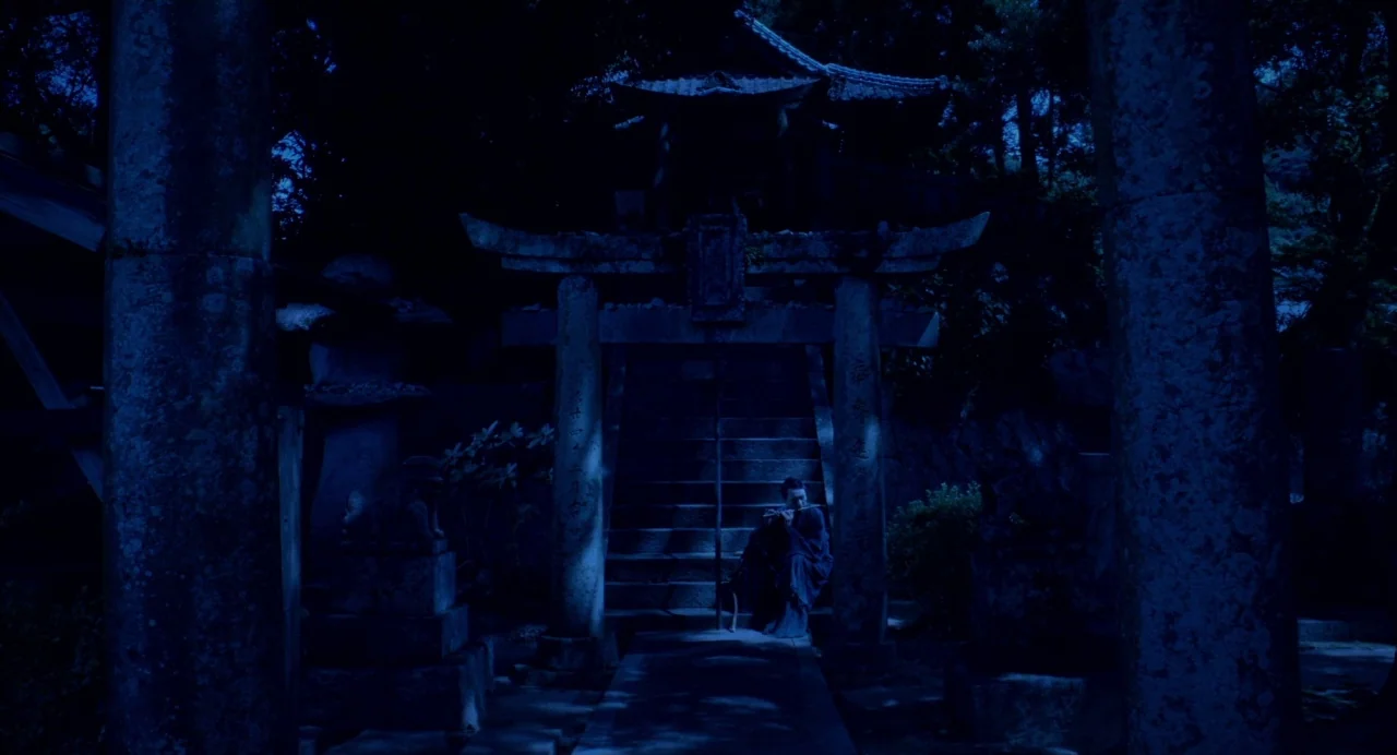 Hanagatami movie still: wide — A traditional stone shrine gate stands at night in a dark wooded area. A man sits on the stone stairs between the…