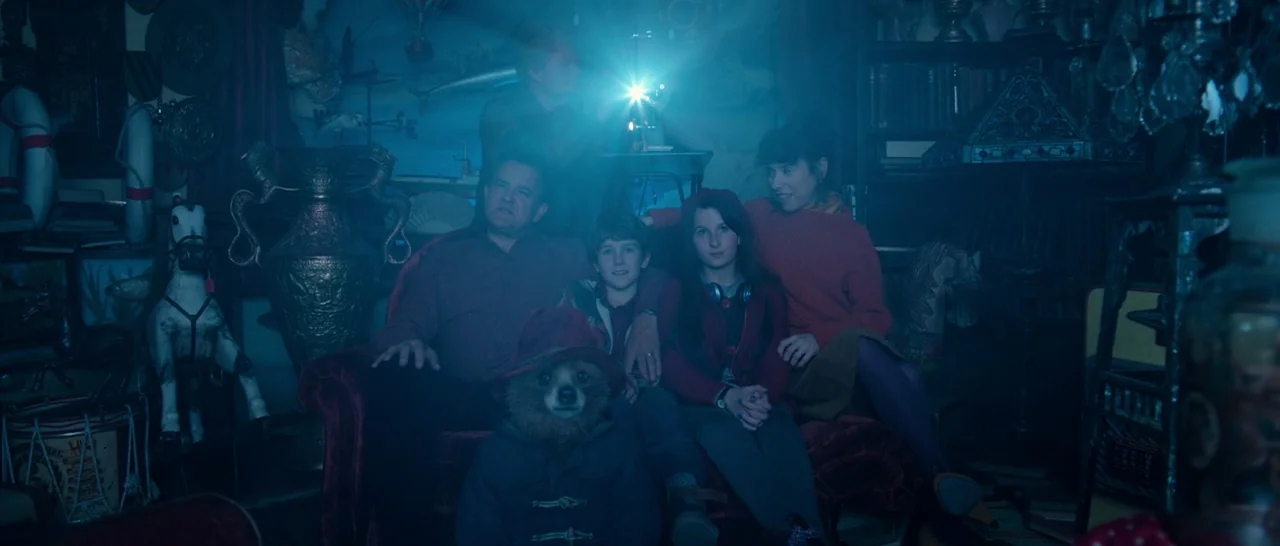 Paddington (2014) movie still: medium — A dimly lit room is filled with antique artifacts and decorative wooden toys. A family of four and a bear…