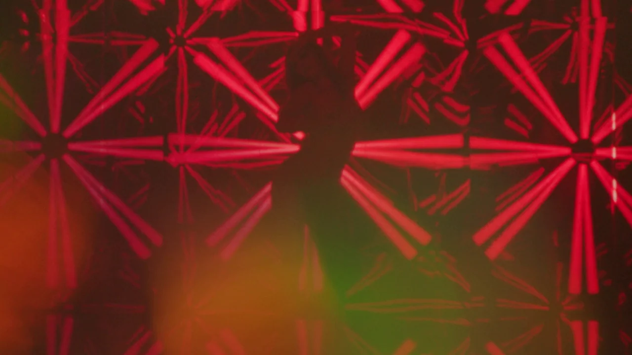Infinity Pool (2023) movie still: medium — A dark, blurred nightclub interior features a wall with intense, repeating red neon patterns. A single…