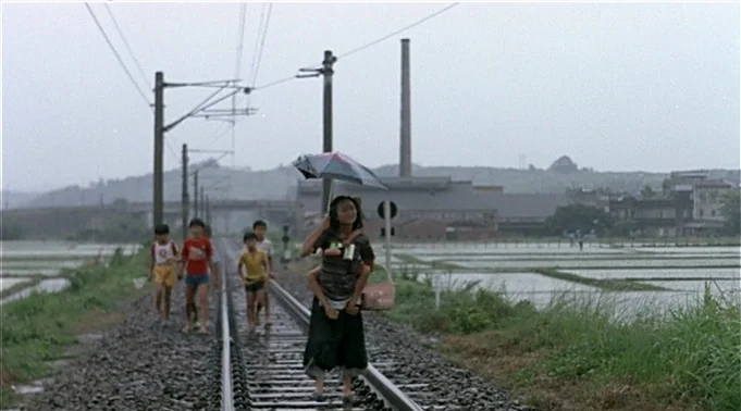 A Summer at Grandpa’s (1984) movie still: wide — A group of children walks along rain-slicked train tracks passing through rural wet paddy fields.…