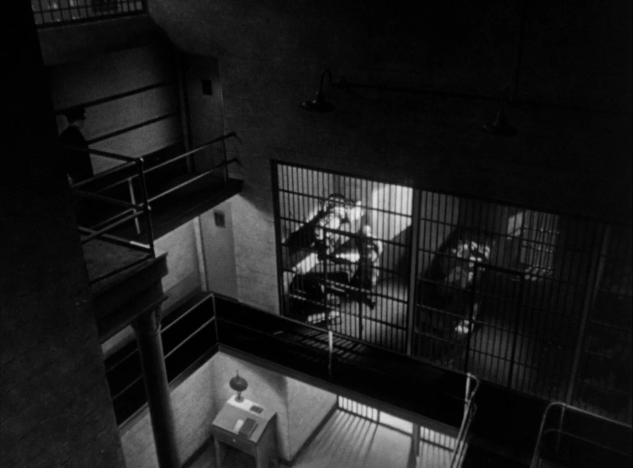 The Killers (1946) movie still: wide — A multi-level prison block features concrete walkways and iron barred doors in the shadows. A desk sits on…