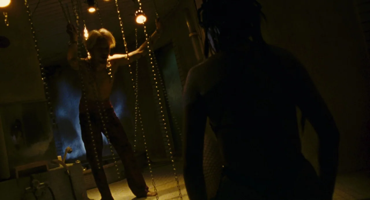 Ichi The Killer (2001) movie still: medium — An industrial-style room features rows of light bulbs dangling from long metal chains. A fair-skinned…