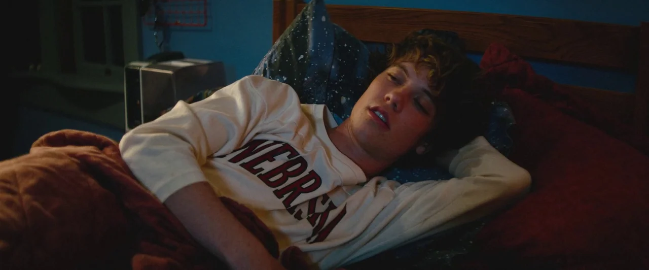 Snack Shack (2024) movie still: medium — A person lies in a bed at night within a dimly lit room. The subject rests with eyes closed and one hand…