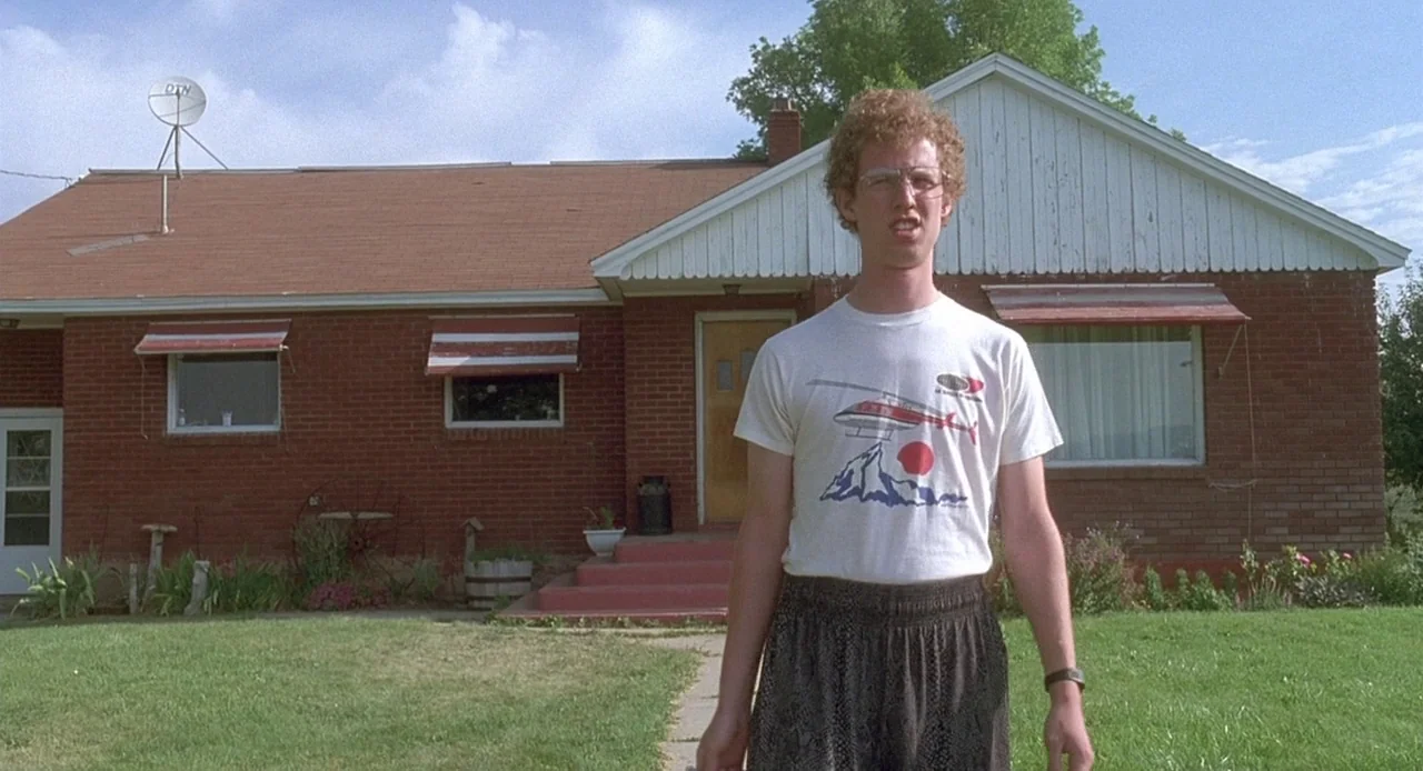 Napoleon Dynamite (2004) movie still: fullbody — A single-story red brick house with a brown roof stands under a blue sky. A young man with curly…