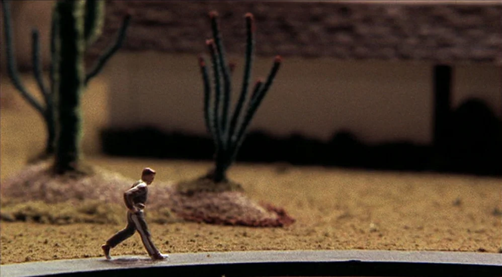 U-Turn (1997) movie still: wide — A miniature desert landscape model features artificial sand, small cactus models, and a building. A small plastic…