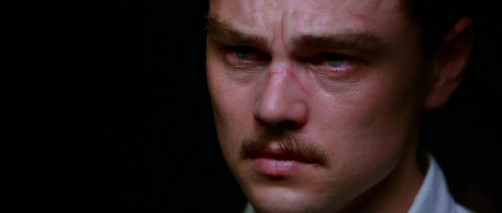 The Aviator (2004) movie still: closeup — A man stands in a dark room with minimal light on his face. He looks forward with a scar visible on the…