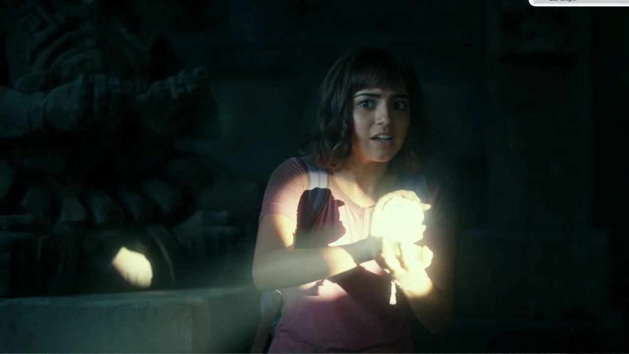 Dora and the Lost City of Gold (2019) movie still: medium — A dark, dimly lit stone chamber contains large carved statues in the background. A young…