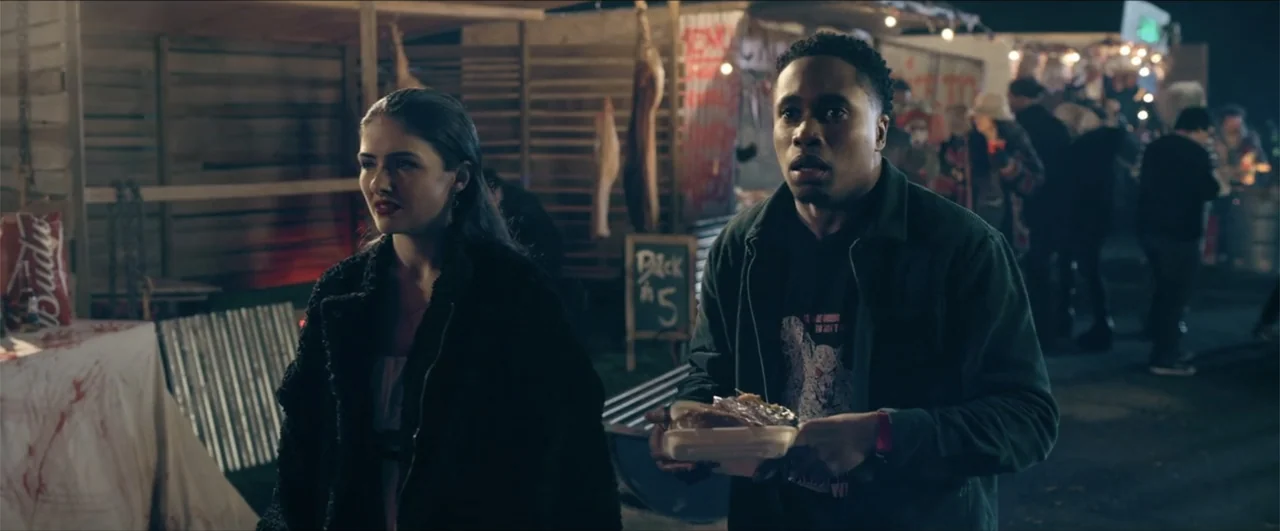 Jeepers Creepers: Reborn (2022) movie still: medium — A night market booth features hanging meat and signs in a dimly lit space. A man and a woman…
