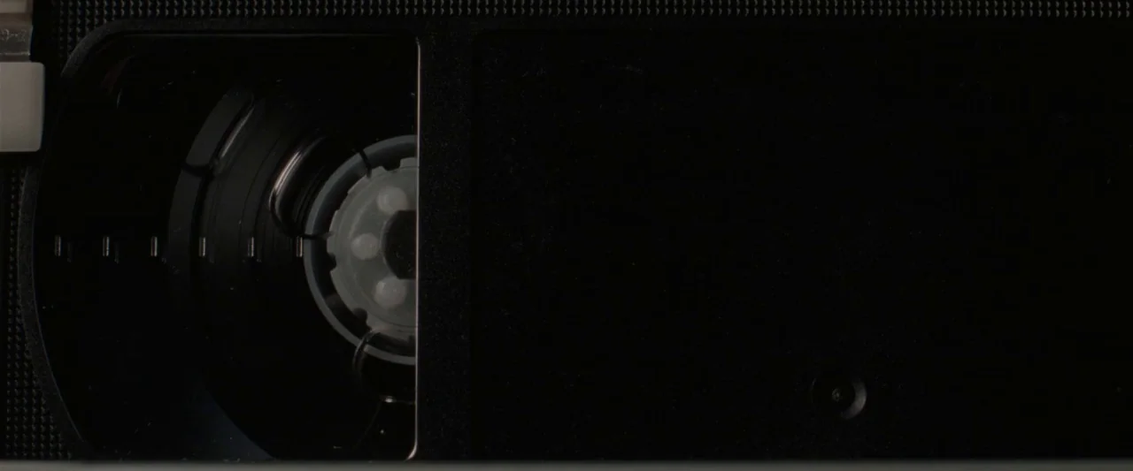 Censor (2021) movie still: closeup — A close-up view of the internal spool mechanism of a black VHS tape. The plastic housing and white inner wheel…