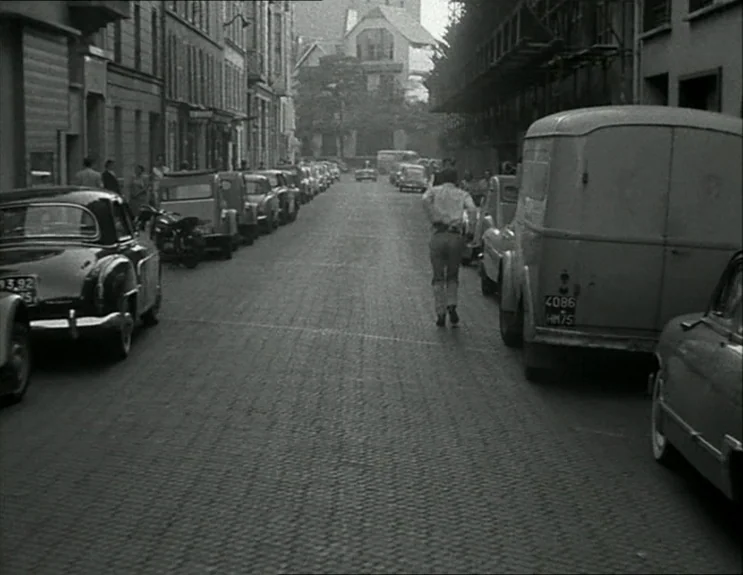 À Bout De Souffle (Breathless) (1960) movie still: fullbody — A narrow cobblestone urban street is lined with parked vintage European automobiles…