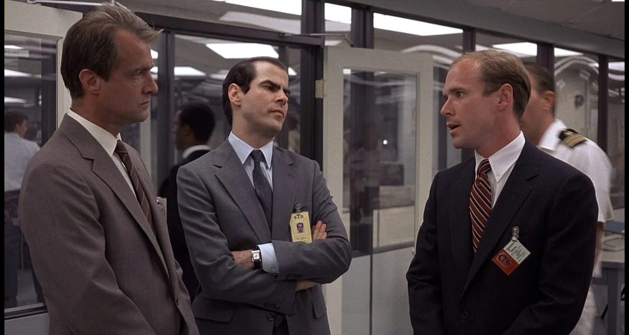 No Way Out (1987) movie still: medium — A sterile corporate office with glass-walled partitions and white suspended ceiling tiles. Three men in…