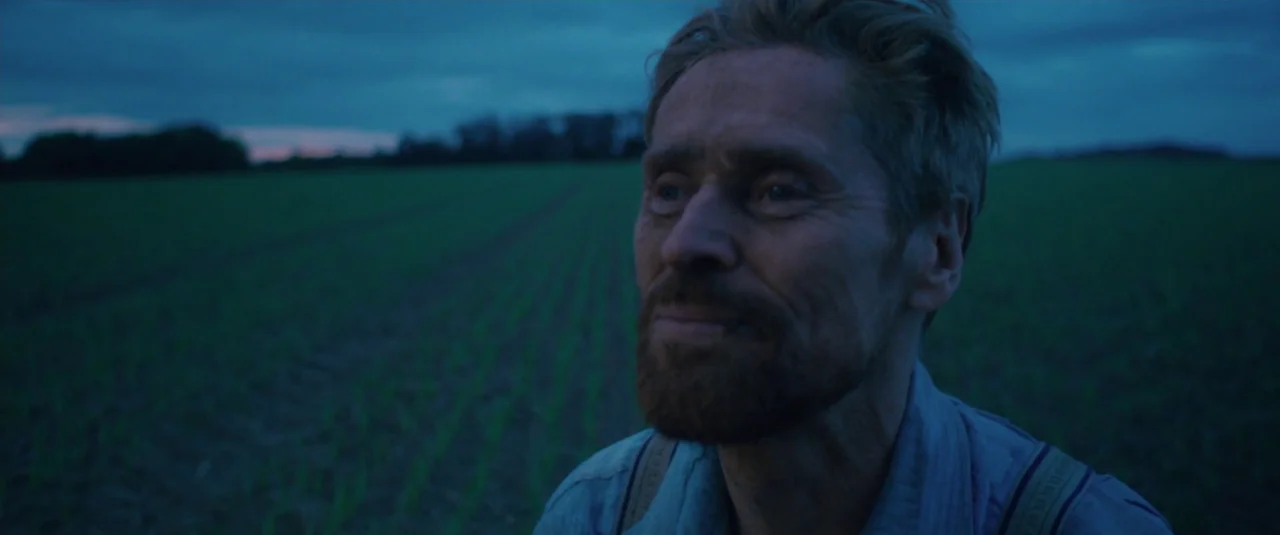 At Eternity’s Gate (2018) movie still: closeup — A vast, open field of young green sprouts sits under a dim blue sky. A man looks off-camera while…