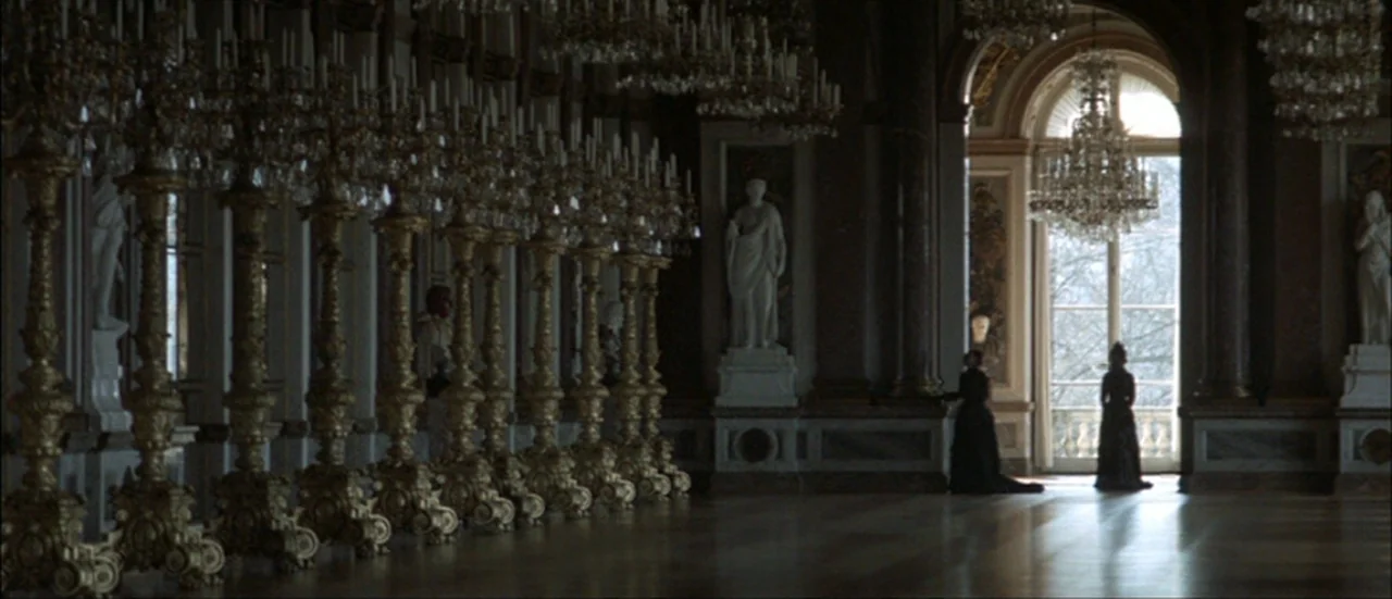 Ludwig (1973) movie still: wide — A long, dimly lit palatial gallery features rows of ornate golden candelabras and statues. Two women in dark…
