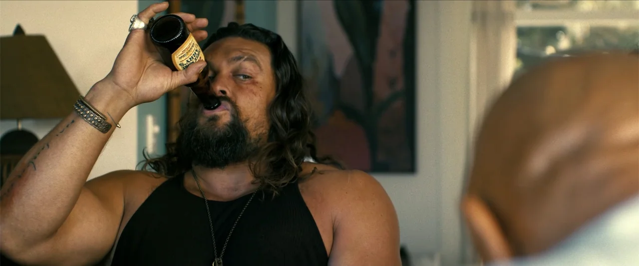 The Wrecking Crew (2026) movie still: medium — A sunlit interior space features a muscular man in a black tank top. The subject holds a brown beer…