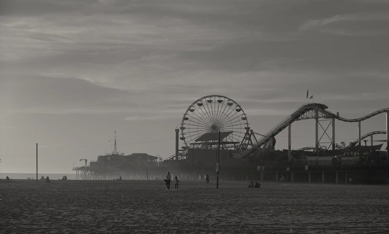 C’mon C’mon (2021) movie still: wide — A wide beach extends beneath an amusement pier featuring a large ferris wheel. Several silhouetted figures…