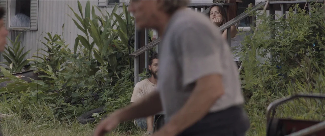 The Peanut Butter Falcon (2019) movie still: medium — A rural mobile home with a wooden porch and overgrown grass in the yard. Three individuals are…