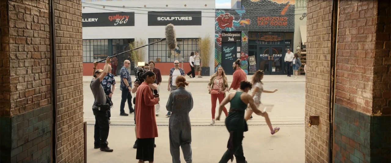 Maintenance Required (2025) movie still: wide — A film production crew works on a set designed to look like a city street. A camera operator records…