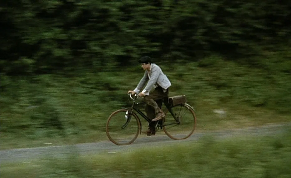 Lacombe, Lucien (1974) movie still: medium — A youth rides a vintage black bicycle along a rural, tree-lined asphalt road. The camera tracks…