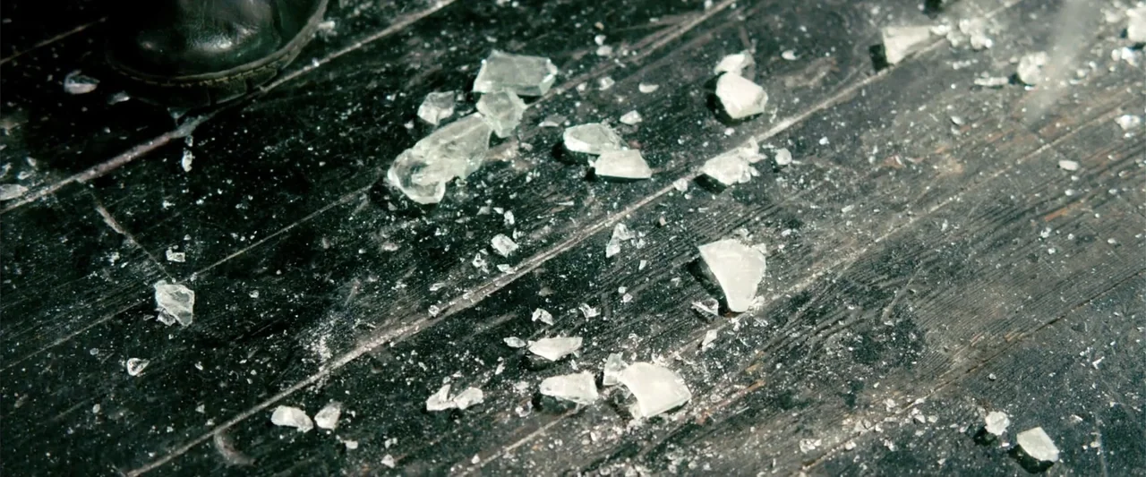 Kick-Ass 2 (2013) movie still: closeup — A dark wooden plank floor is covered with scattered pieces of broken clear glass. A partial view of a black…