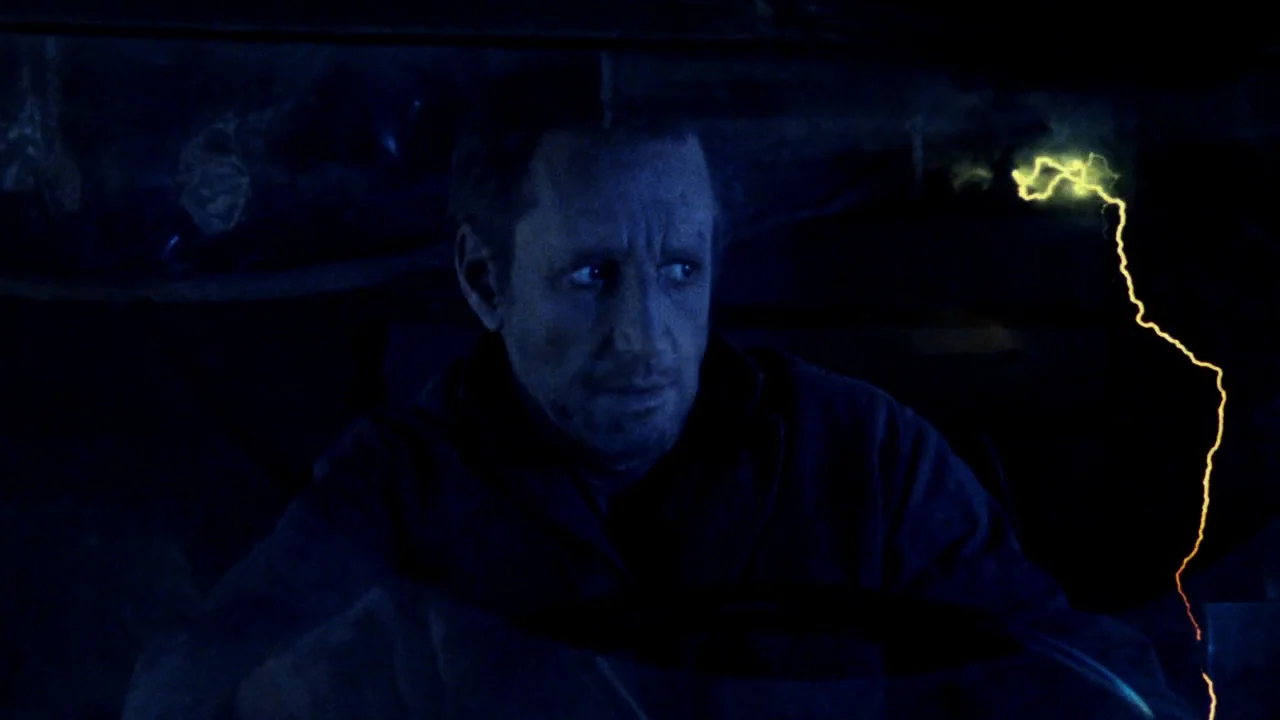 Sorcerer (1977) movie still: closeup — The interior of a vehicle is dark with only faint illumination. A man is driving while looking toward the…