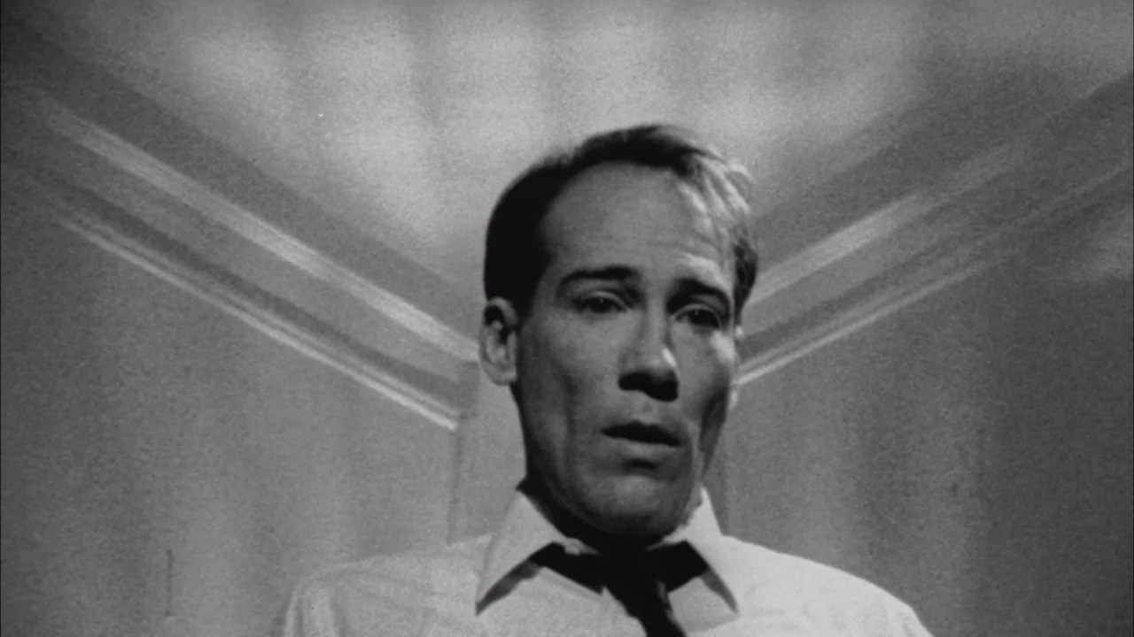 Poison movie still: closeup — A man in a collared shirt stands against a light-colored corner wall. He gazes forward while harsh lighting from above…