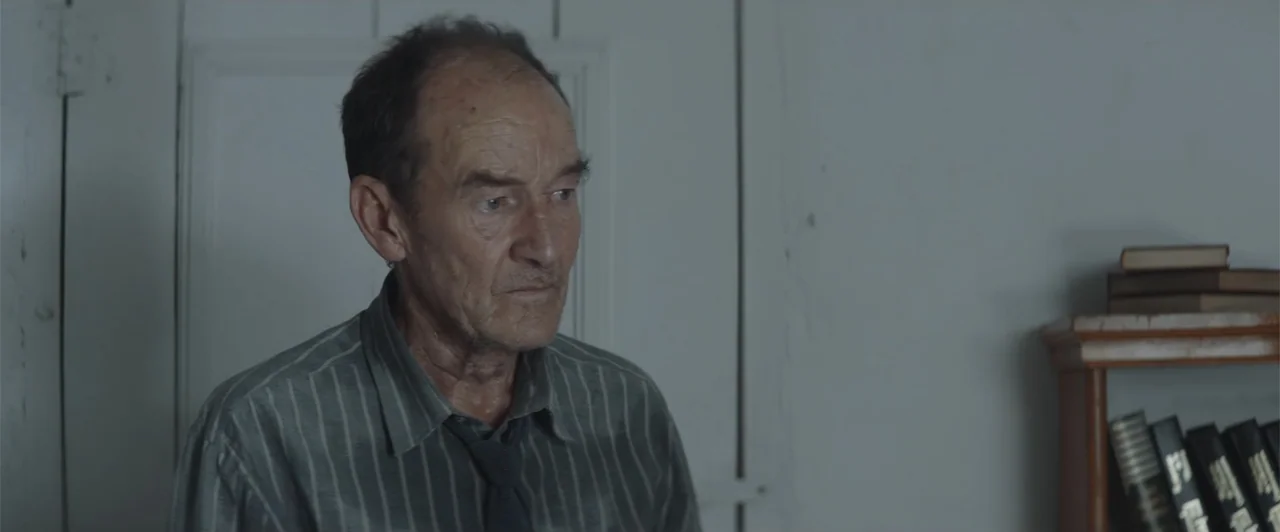 My Neighbor Adolf (2022) movie still: medium — A spartan study contains a wooden bookshelf filled with several hardbound books. An elderly man in a…