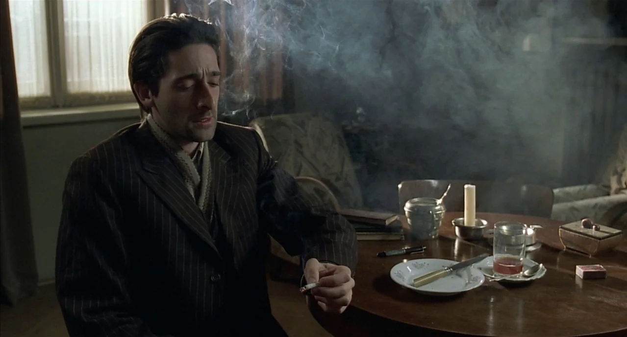 The Pianist (2002) movie still: medium — A dimly lit room features a round wooden table scattered with remnants of a meal. A man in a pinstripe suit…