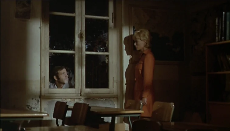 Le Boucher (1970) movie still: medium — A dimly lit classroom at night features rows of wooden desks and chairs. A man peers through the glass…
