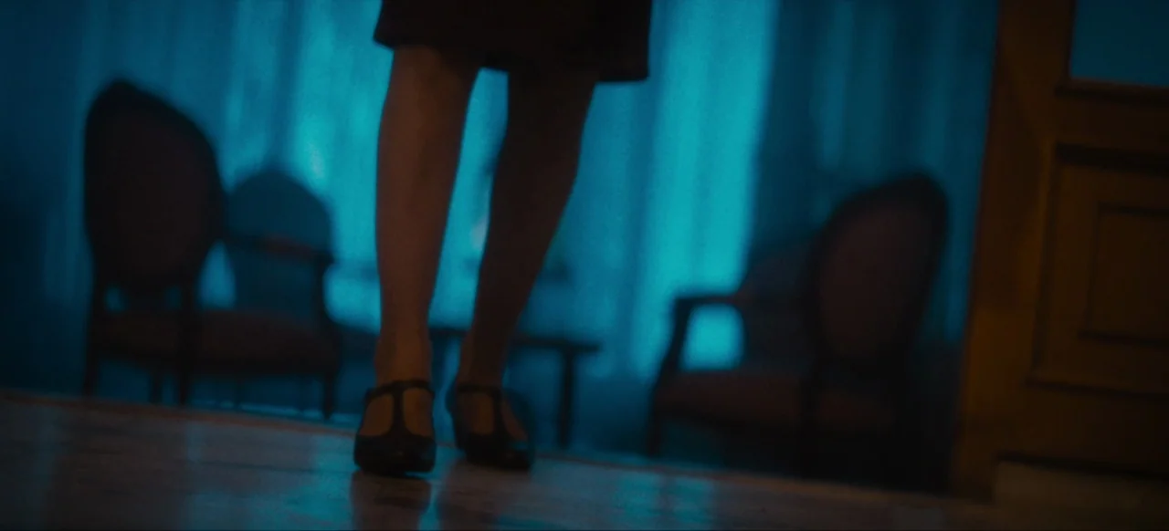 Housewife (2017) movie still: medium — A dimly lit room with two chairs flanking a window with blue-lit curtains. The subject stands on a wooden…